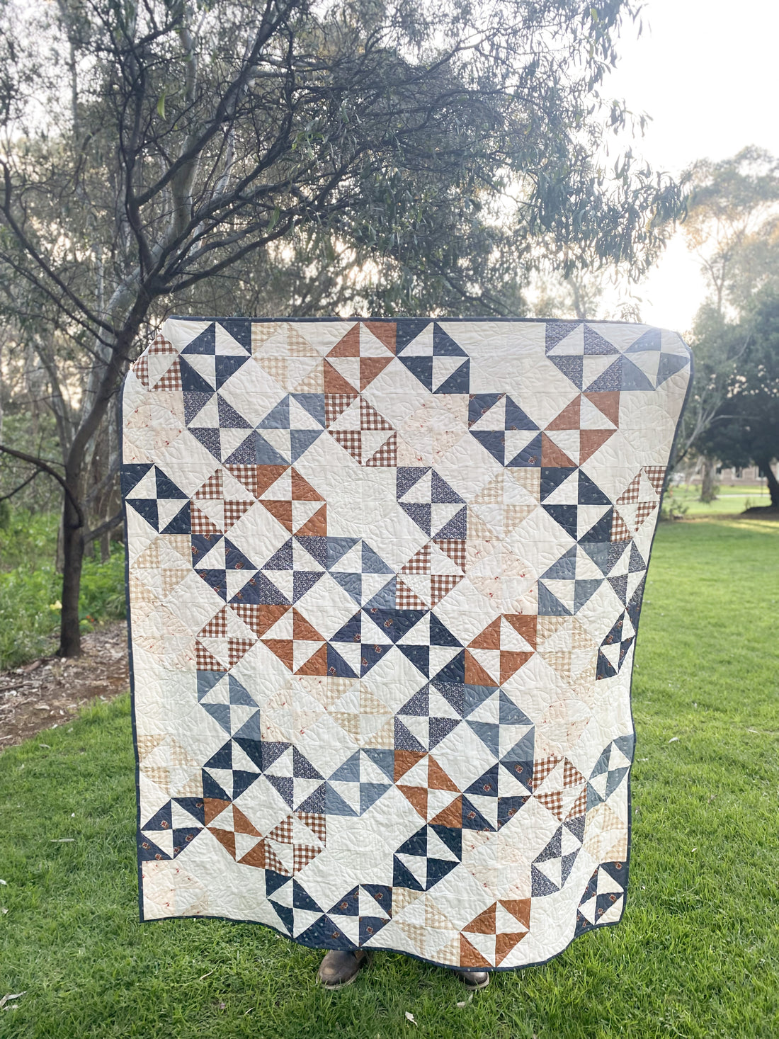Quilt  with cream, blue and brown fabrics, held up in park with trees